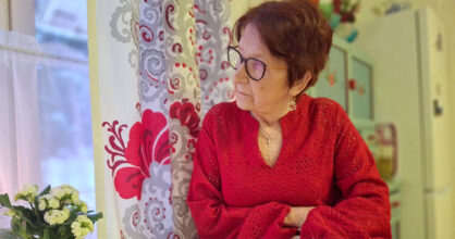 Iäkäs silmälasipäinen nainen katsoo ikkunasta ulos.