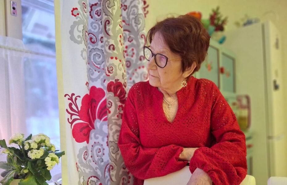 Iäkäs silmälasipäinen nainen katsoo ikkunasta ulos.