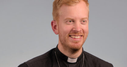 Mies hymyilee. Hänellä on papin paita päällä. Hänen hiuksensa ovat oranssinväriset.