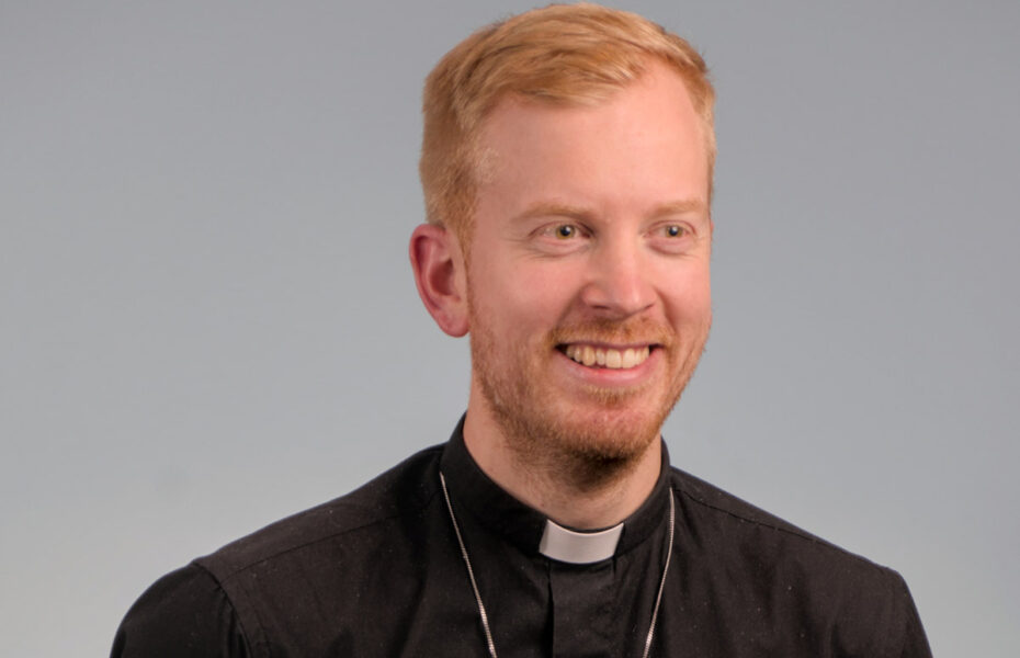 Mies hymyilee. Hänellä on papin paita päällä. Hänen hiuksensa ovat oranssinväriset.