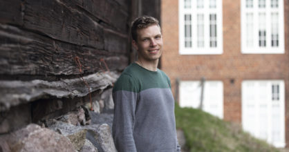 Matias Saarenpää seisoo vanhan puurakennuksen edustalla. Hän hymyilee yllään collegepaita.