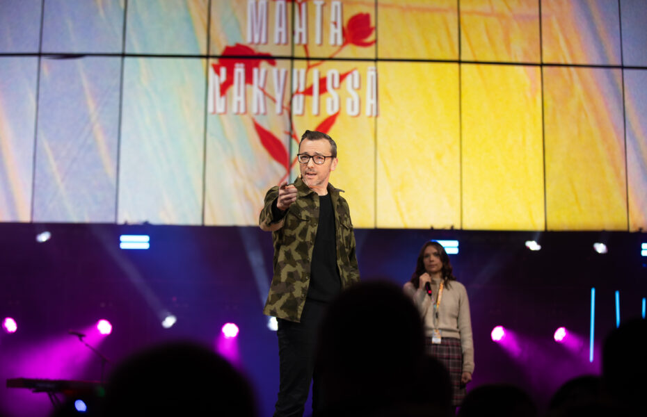 Silmälasipäinen tummahiuksinen mies osoittaa sormellaan yleisöön rohkaisevasti.