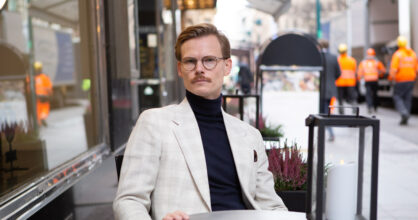 Mies istuu kahvilan edessä valkoinen takki ja poolokaulus paita yllään.