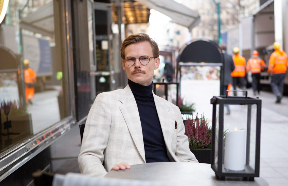 Mies istuu kahvilan edessä valkoinen takki ja poolokaulus paita yllään.