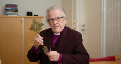 Papinpaitaan pukeutunut harmaantunut ja silmälasipäinen mies pitelee käsissään koristeellista ristiä pöydän ääressä.