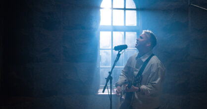 Mies soittaa kitaraa ja laulaa hämärässä huoneessa. Valo loistaa ikkunasta.