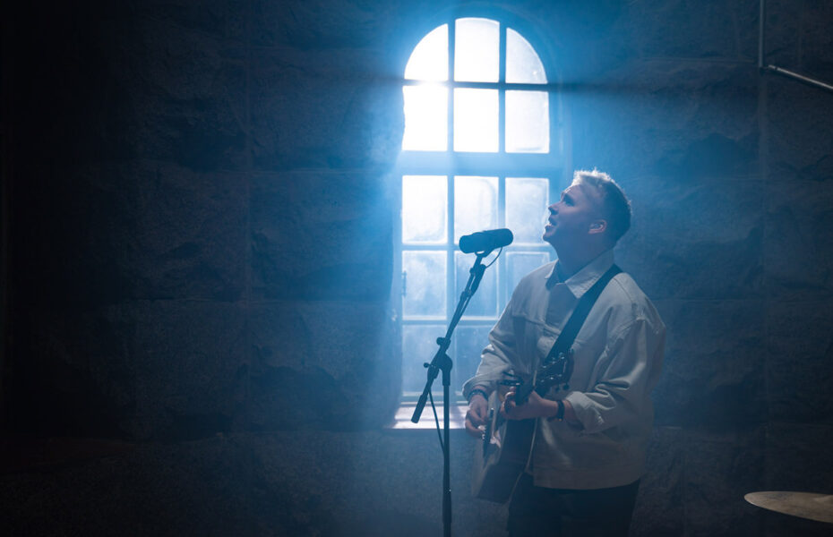 Mies soittaa kitaraa ja laulaa hämärässä huoneessa. Valo loistaa ikkunasta.