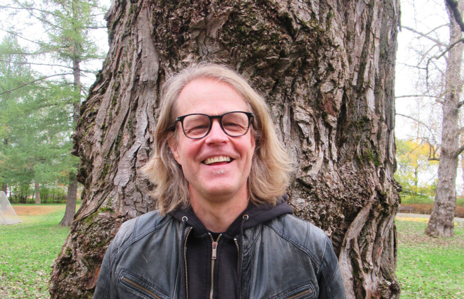 Mikko Miettinen seisoo puun edessä. Hänellä on vaaleat hiukset, jotka ulottuvat hartioille, nahkatakki ja silmälasit.