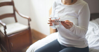 Nainen istuu sängyn reunalla ja pitää kädessää vesilasia ja pillereitä.