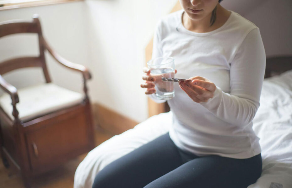 Nainen istuu sängyn reunalla ja pitää kädessää vesilasia ja pillereitä.