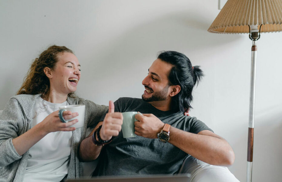 Nainen ja mies istuvat sohvalla kupit käsissään. He nauravat.