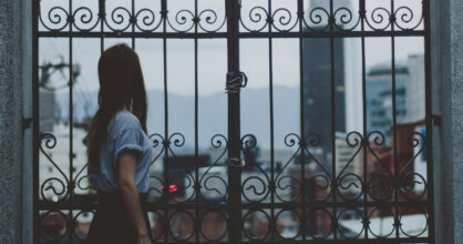 Nainen seisoo lukitun portin takana. Portin takana on harmaa kaupunki.