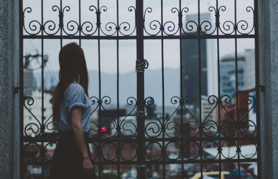 Nainen seisoo lukitun portin takana. Portin takana on harmaa kaupunki.