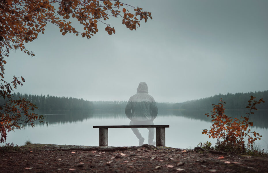 Näkymätön mies istuu penkillä järven rannalla.