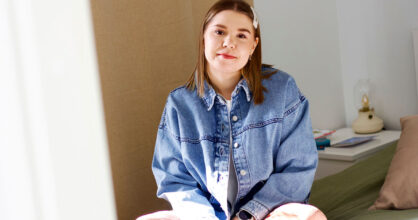 Nainen istuu sängyllä. Hänellä on farkkutakki.