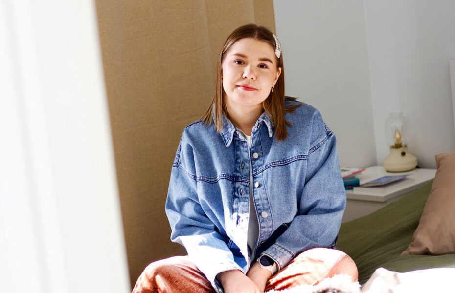 Nainen istuu sängyllä. Hänellä on farkkutakki.