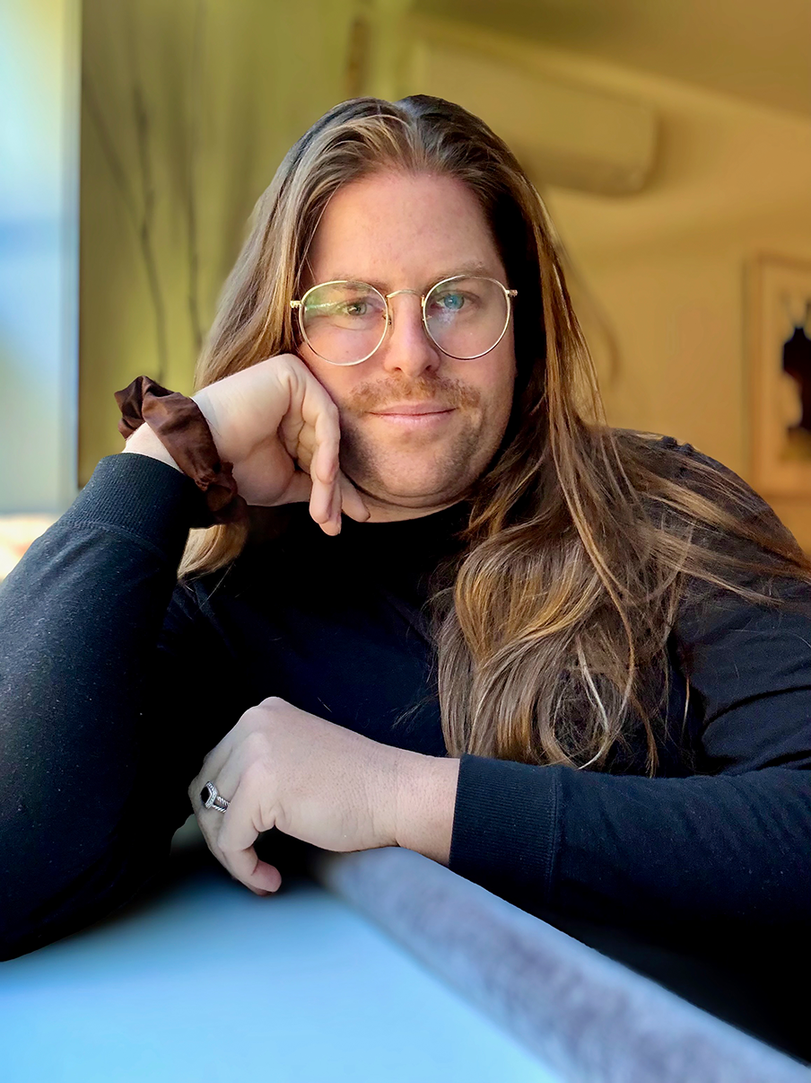 Mies nojaa käteensä ja katsoo kohtisuoraan. Hänellä on pitkät ruskeat hiukset ja silmälasit, sekä lyhyt parta.