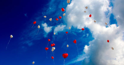 Sydämenmuotoisia ilmapalloja lentää taivaalle. Taivaalla on pilviä.