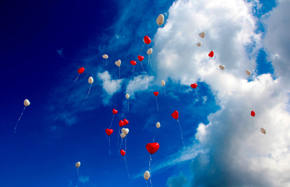 Sydämenmuotoisia ilmapalloja lentää taivaalle. Taivaalla on pilviä.