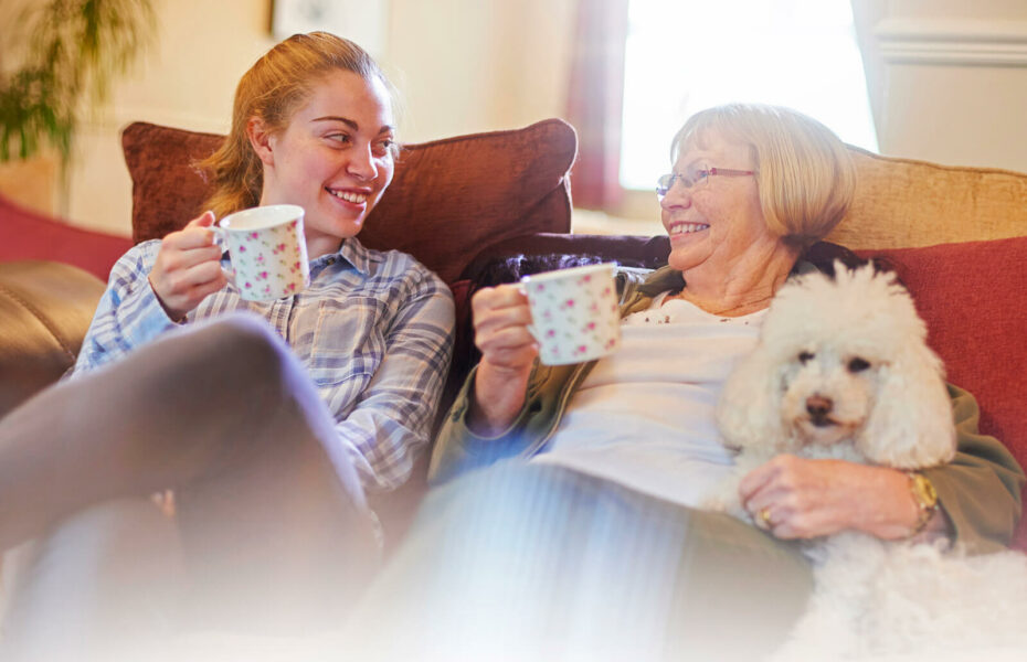 Kuvassa nuori ja vanha nainen istuvat sohvalla hymyillen ja pidellen kahvikuppeja.
