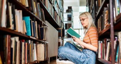 Nuori nainen istuu lattialla kirjahyllyjen välissä ja lukee kirjaa.