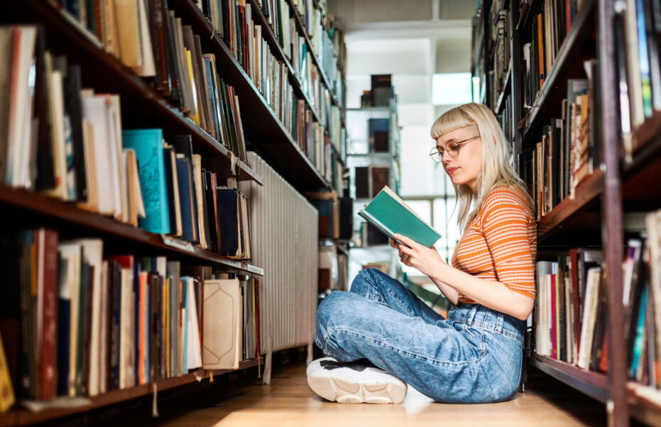 Nuori nainen istuu lattialla kirjahyllyjen välissä ja lukee kirjaa.