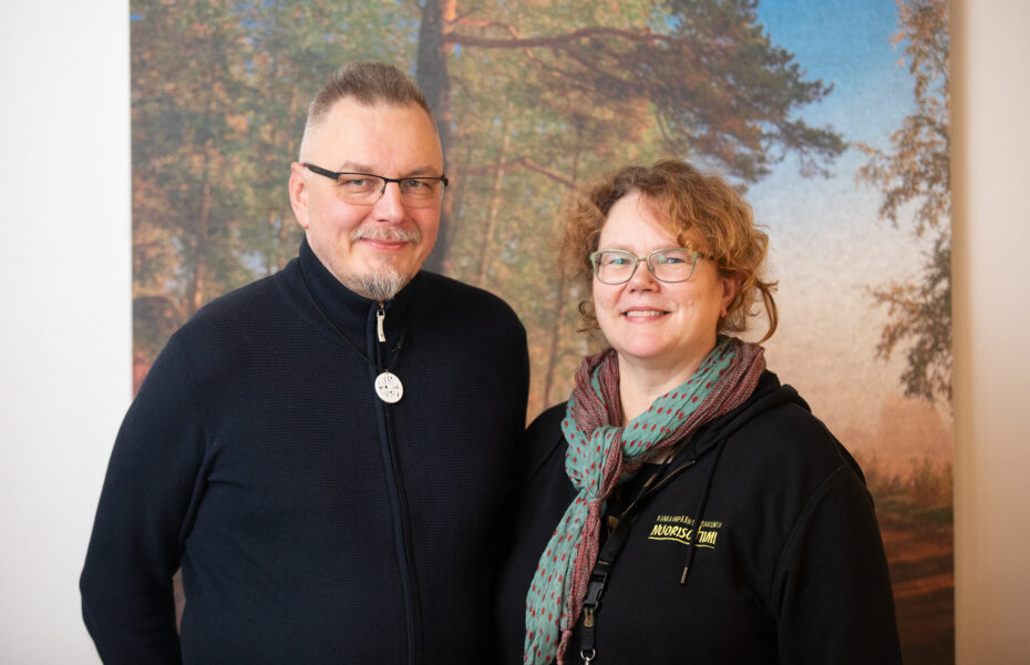 Mies ja nainen hymyilevät sisällä. Naisella lukee hupparissa "nuorisotiimi".