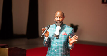 Pitkäpartainen mies puhumassa lavalla mikkiin.