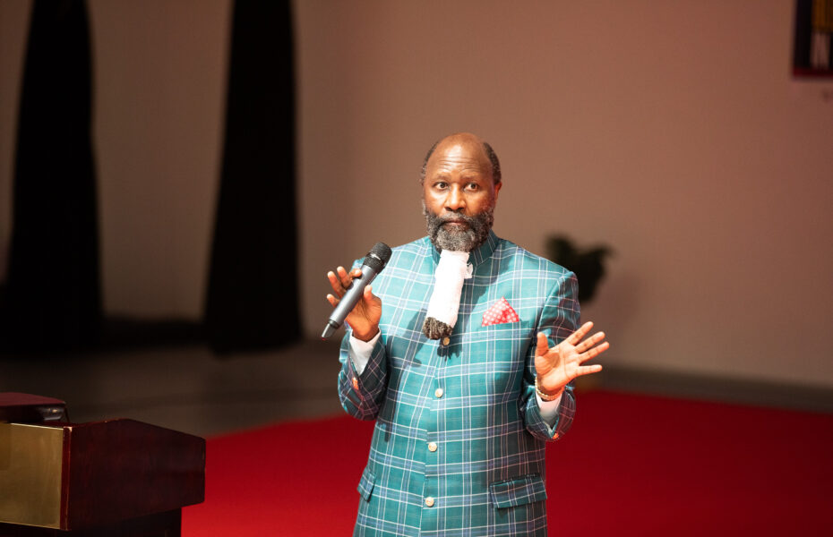 Pitkäpartainen mies puhumassa lavalla mikkiin.