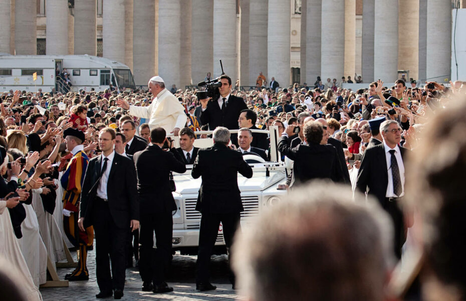 Paavi Franciscus tervehtii ihmisiä autostaan. Häntä ympäröivät turvamiehet.