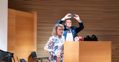 Päivi Räsänen seisoo puhujanpöntön takana. Hänen takanaan Niilo Räsänen ottaa kännykällä valokuvaa.