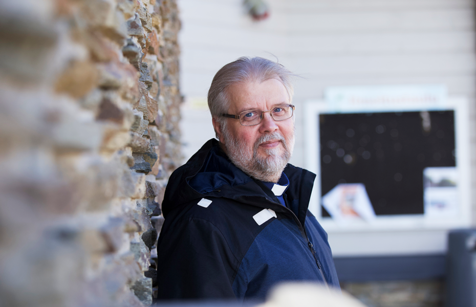 Silmälasipäinen vanhempi mies hymyilee ulkona. Hänellä on papin "sokeripala".