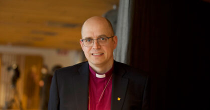 Kalju ja silmälasipäinen mies hymyilee. Hänellä on musta puvun takki ja sen alla violetti papinpaita.