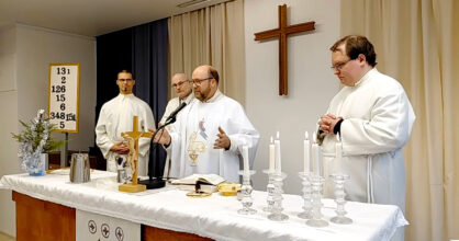 Pastori seisoo valkoisessa albassa alttarin takana, vierellään kolme ehtoollisavustajaa