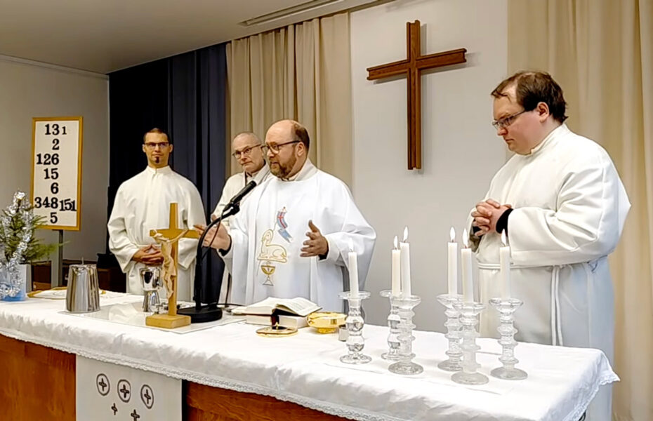 Pastori seisoo valkoisessa albassa alttarin takana, vierellään kolme ehtoollisavustajaa