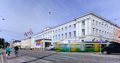 Helsingin valtioneuvoston linnan edustalla liehuu sateenkaariliput. Talon edestä menee raitiovaunu.