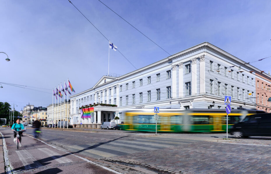 Helsingin valtioneuvoston linnan edustalla liehuu sateenkaariliput. Talon edestä menee raitiovaunu.