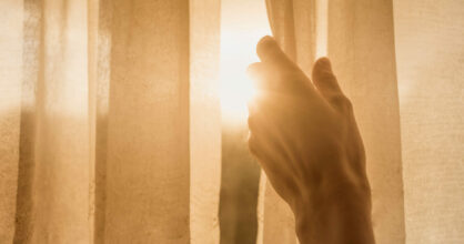 Kuvassa käsi raottaa ikkunaverhoa, mistä kajastaa auringonvalo.