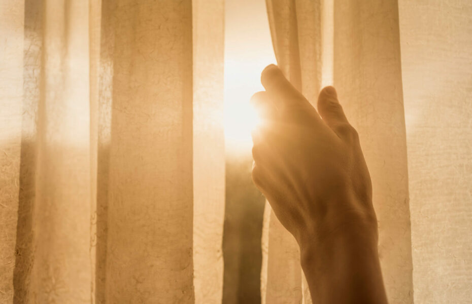 Kuvassa käsi raottaa ikkunaverhoa, mistä kajastaa auringonvalo.