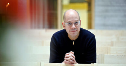 Mies istuu penkkirivillä. Hän nojaa käsiinsä, jotka ovat ristissä ja katsoo hymyillen.