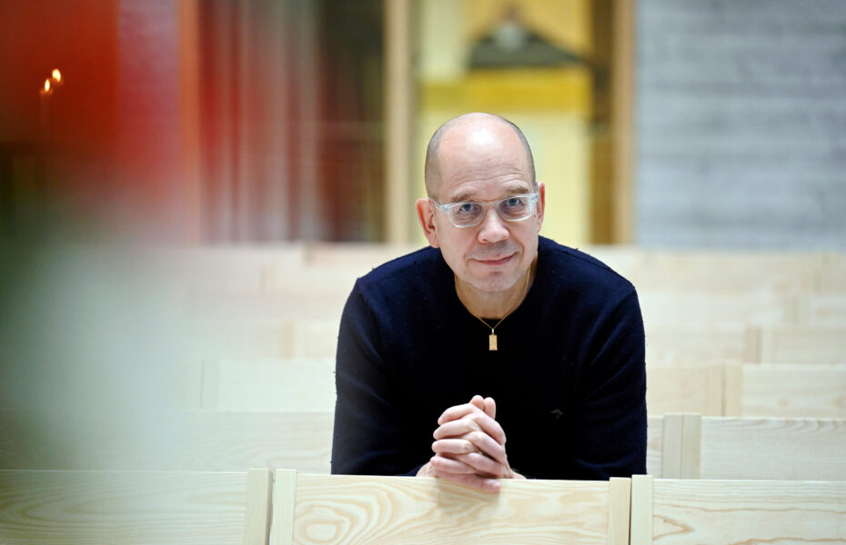 Mies istuu penkkirivillä. Hän nojaa käsiinsä, jotka ovat ristissä ja katsoo hymyillen.