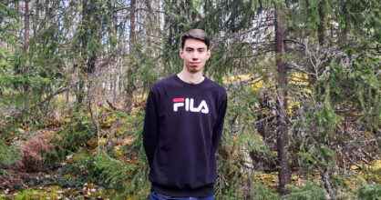 Nuori mies seisoo kuusimetsän laidalla ja hymyilee.