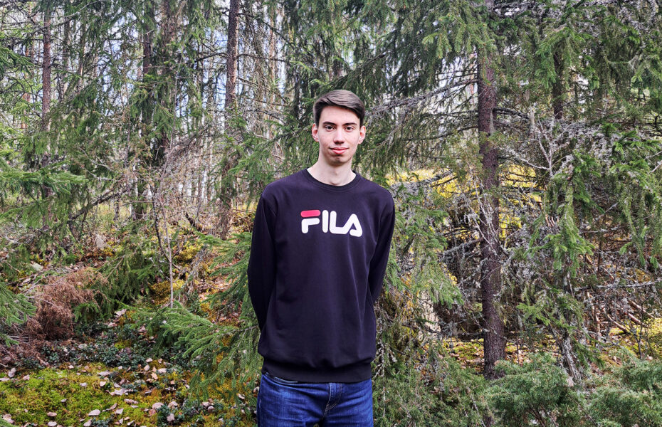 Nuori mies seisoo kuusimetsän laidalla ja hymyilee.