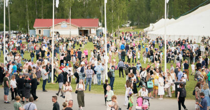 Paljon ihmisiä ruohikkoisella alueella, jonka etualalla menee tie. Taustalla kaksi isoa telttarakennusta.