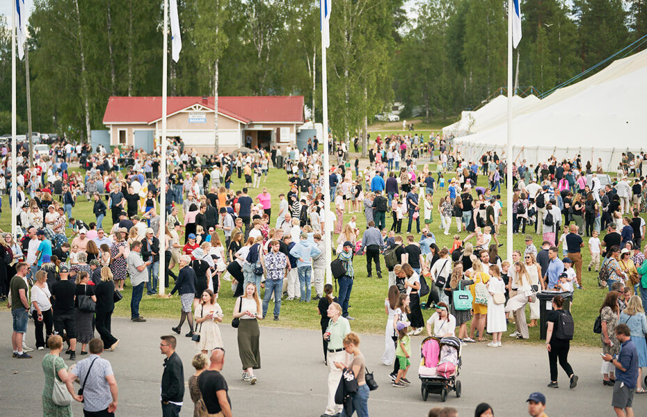 Paljon ihmisiä ruohikkoisella alueella, jonka etualalla menee tie. Taustalla kaksi isoa telttarakennusta.