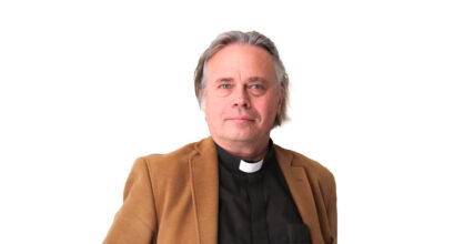 Harmaahiuksisella miehellä on papinpaita ja ruskea takki.