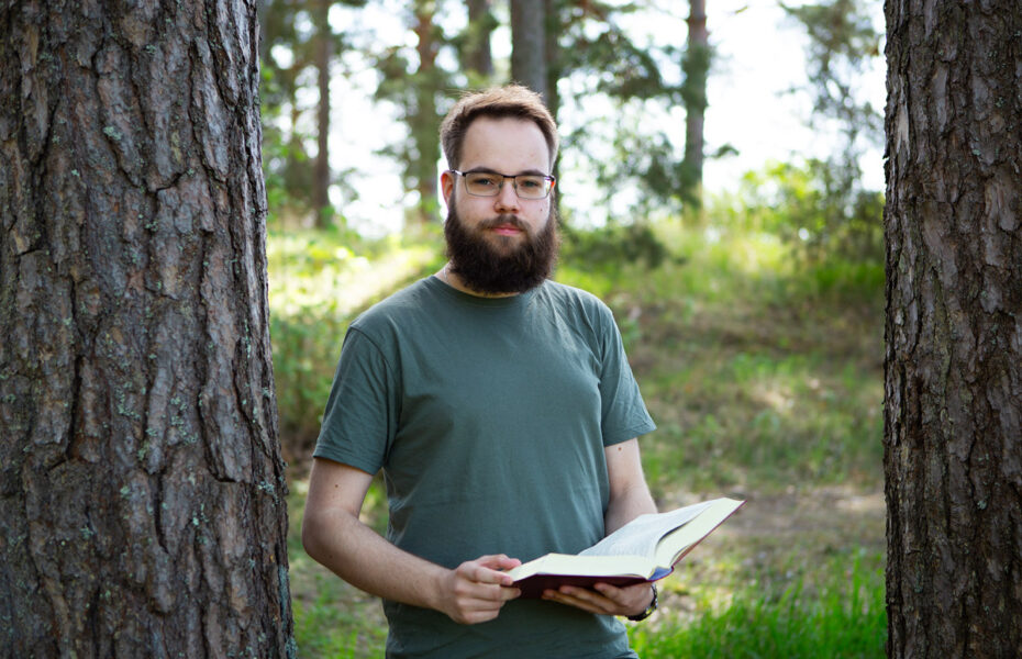 Parrakas nuorimies seisoo kahden puun välissä ja pitää käsissään kirjaa.