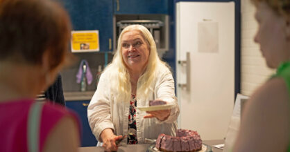 Seija Nurmi tarjoaa pöydän yli laustasta, jossa on kakun palanen. Hänellä on vaaleat pitkät hiukset.