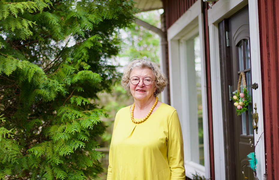 Silmälasipäinen vanhempi nainen keltaisessa paidassa. Ulkona. Taustalla puu ja talo.