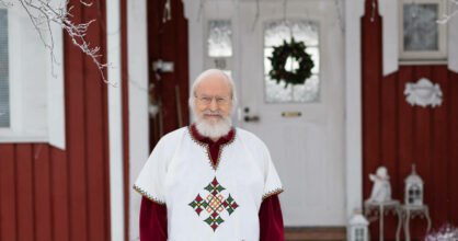 Seppo Väisänen seisoo omakotitalon ulko-oven edessä. Hänellä on valkoinen parta ja hiukset sekä silmälasit.